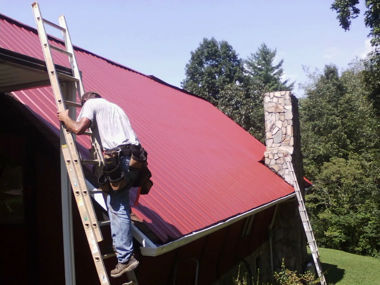 Roofer on a ladder beside a red metal roof and stone chimney — Soffit & Fascia Repair by Roofing Brothers in Durkee